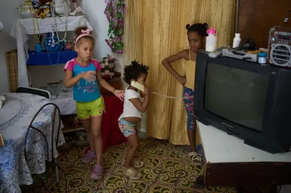 ثلاث فتيات صغيرات يلعبن في غرفة بسيطة في هافانا، مع تلفاز قديم وأثاث متواضع، تعكس صورة الحياة اليومية في ظل الأزمة الاقتصادية.