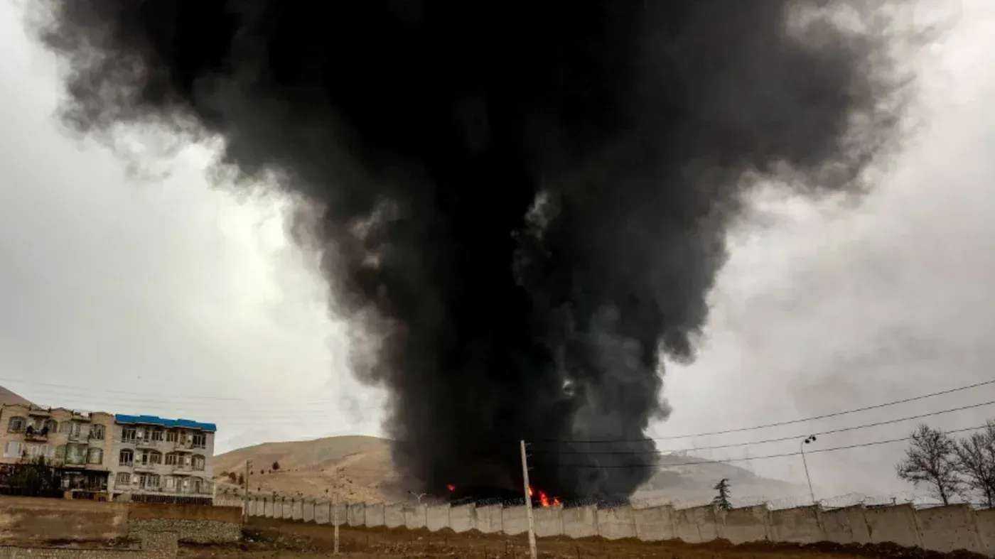 دخان أسود كثيف يتصاعد من موقع تعرض لهجوم، مع وجود مبانٍ قريبة في الخلفية، مما يعكس تصاعد التوترات في الصراع الإيراني.