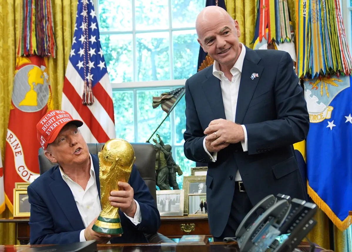 رئيس FIFA جياني إنفانتينو يقف مبتسمًا بجانب دونالد ترامب الجالس ويحمل كأس العالم في مكتب رسمي مع أعلام خلفهما، في سياق مناقشة خطة بيع عائدات كأس العالم.