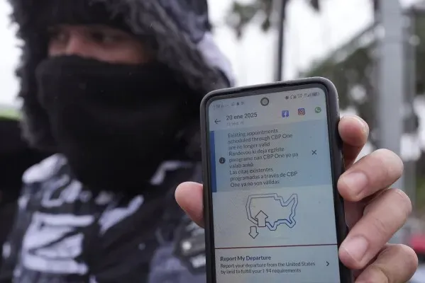 شخص يرتدي ملابس دافئة يحمل هاتفًا يظهر إشعارًا متعلقًا بتطبيق "CBP One" الخاص بجدولة المواعيد للعبور إلى الولايات المتحدة.