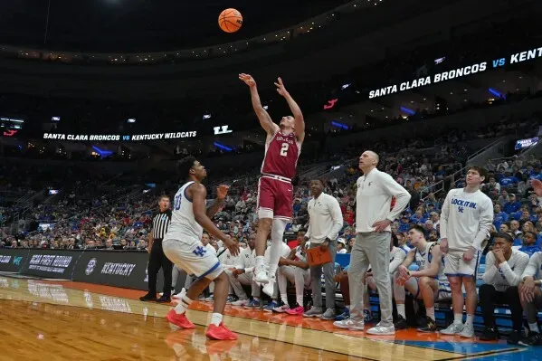 لاعب سانتا كلارا يسدد كرة سلة أثناء مباراة ضد كنتاكي، مع تواجد لاعبي الفريقين والمدرب على الجانبين في أجواء تنافسية حماسية.