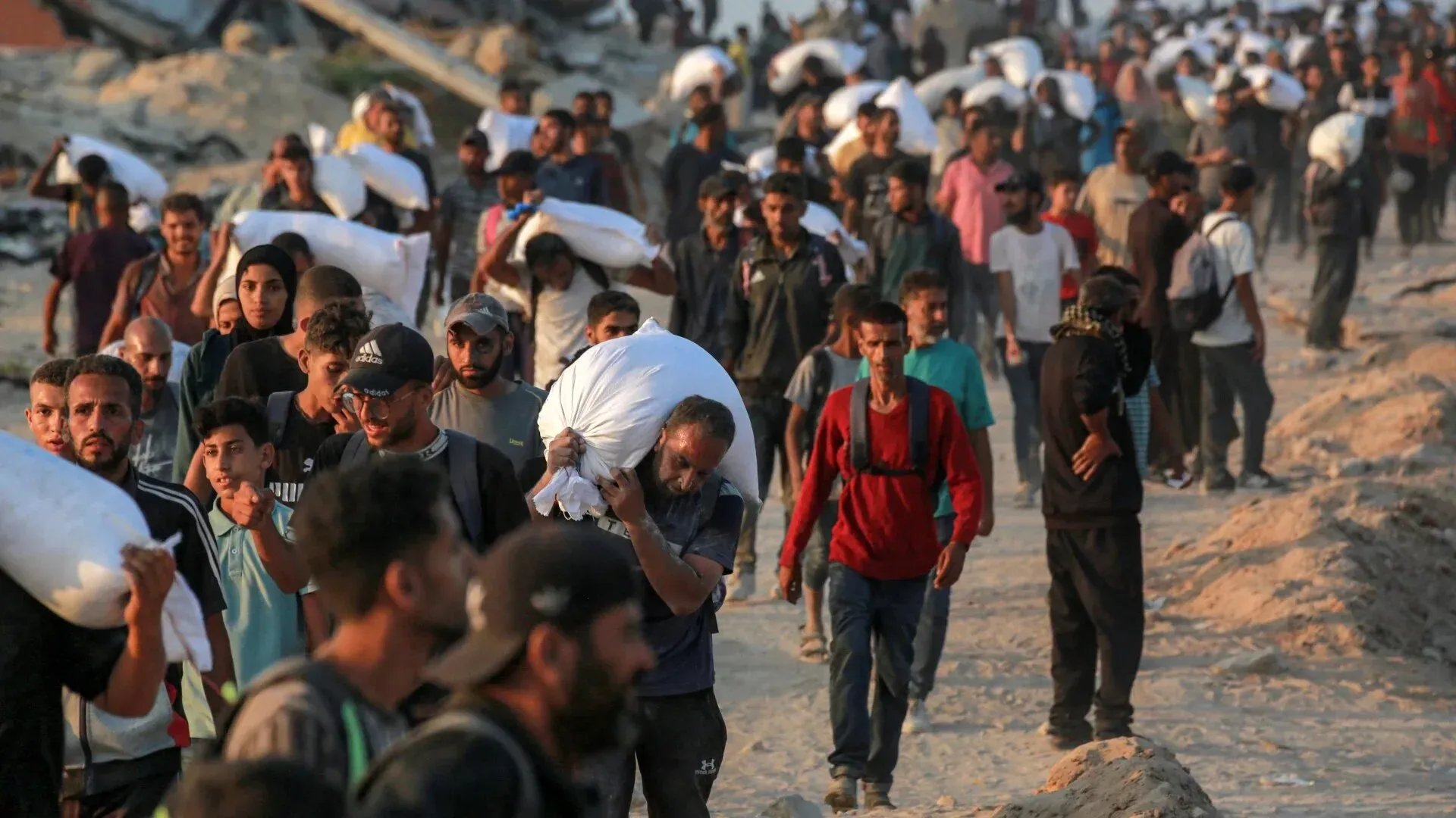 مئات الفلسطينيين في غزة يحملون أكياس المساعدات الغذائية، في ظل ظروف خطيرة وقيود مشددة، بحثًا عن الغذاء وسط الصراع.