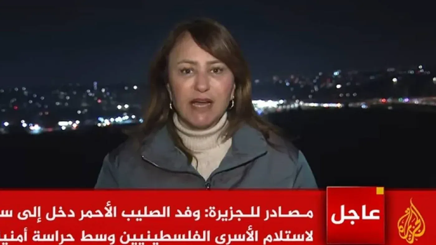 صحفية من قناة الجزيرة تغطي أحداث الإفراج عن الأسرى الفلسطينيين، مع وجود خلفية مضاءة لمدينة فلسطينية في الليل.