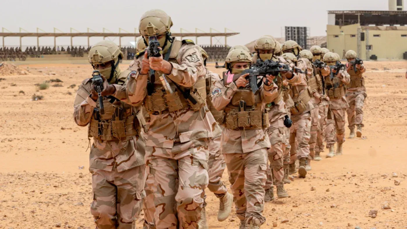 جنود يرتدون زيًا عسكريًا كاملًا، يحملون أسلحة، يسيرون في صفوف منتظمة في منطقة صحراوية، مما يعكس تدريبات عسكرية محتملة لدعم قوات حفتر في ليبيا.