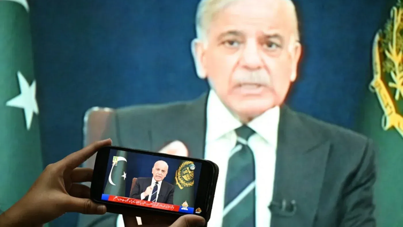 رجل يتحدث في مؤتمر صحفي، يظهر على شاشة كبيرة، بينما يُسجل شخص ما حديثه بهاتفه المحمول، مع العلم الوطني الباكستاني في الخلفية.