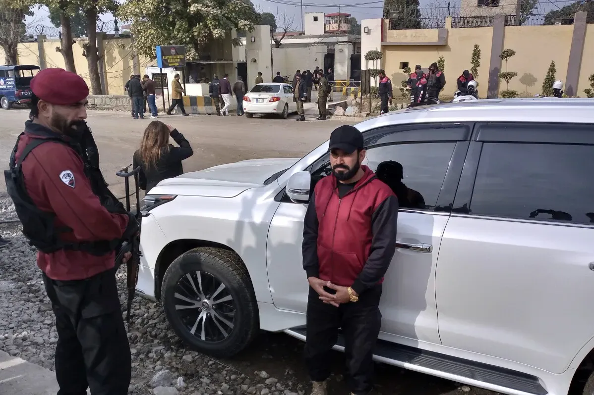 حارس مسلح يقف بجانب سيارة بيضاء خارج سجن أديالا، بينما تتجمع مجموعة من الناس في الخلف، في سياق حكم السجن لعمران خان وزوجته.