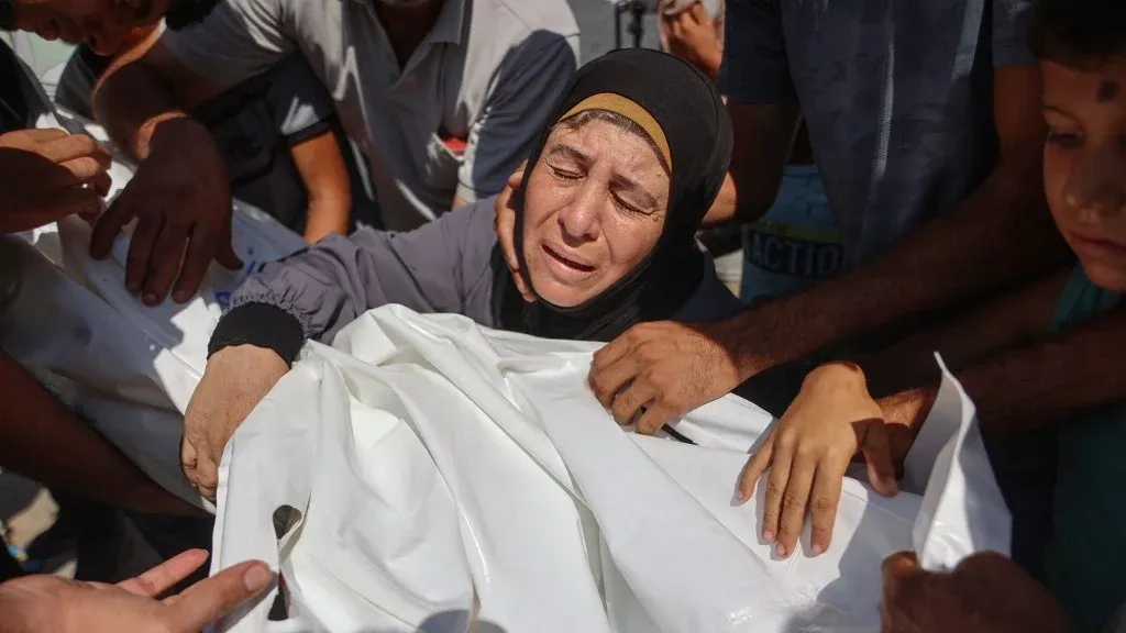امرأة فلسطينية تبكي وهي تحيط بجثمان ملفوف، تعبير عن الألم والخسائر في غزة وسط استمرار الإبادة الجماعية والهجمات الإسرائيلية.