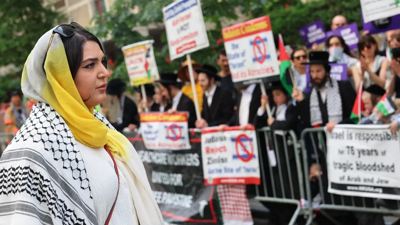 ناشطة فلسطينية-أمريكية ترتدي حجابًا وتظهر في مظاهرة، محاطة بلافتات تعبر عن دعم فلسطين، تعكس التوترات حول حقوق الفلسطينيين.
