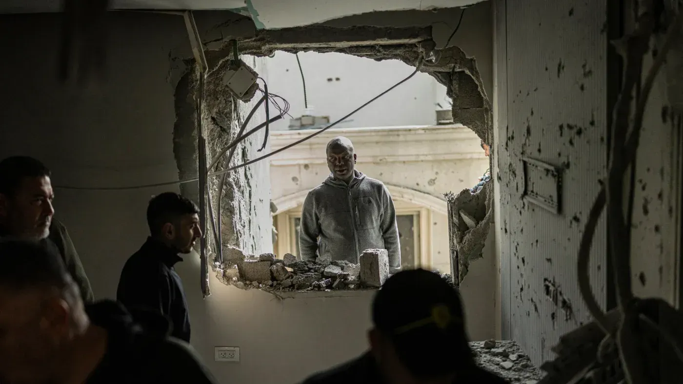 شخص ينظر من خلال فتحة في جدار مدمّر، بينما يتجمع آخرون في الداخل، مما يعكس آثار الصراع والدمار في المجتمعات الفلسطينية.