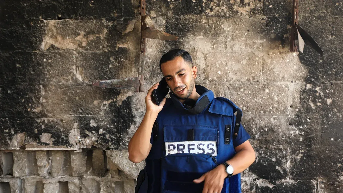 صحفي فلسطيني يرتدي سترة "صحافة" ويتحدث عبر الهاتف، يقف أمام جدار متضرر، يعكس ظروف العمل الصعبة في غزة.
