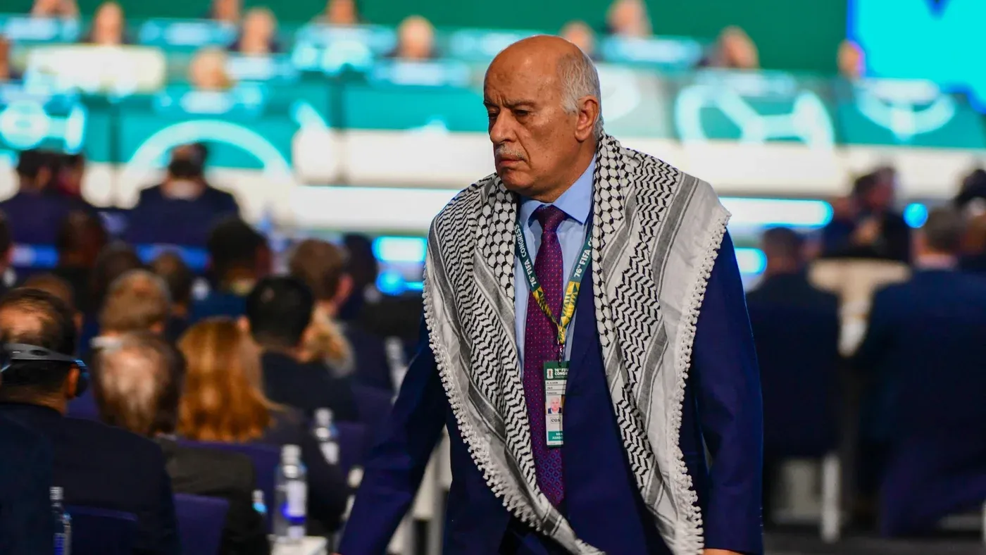 جبريل رجوب، رئيس الاتحاد الفلسطيني لكرة القدم، يرتدي الكوفية ويظهر في مؤتمر، وسط حشد من الحضور، مع التركيز على قضايا التأشيرات والمشاركة في كأس العالم.