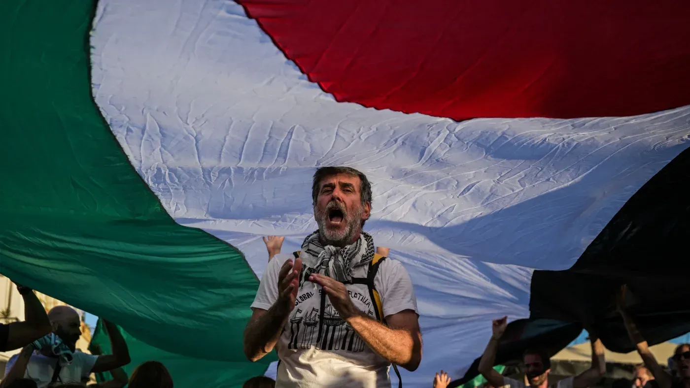 رجل يحمل علم فلسطين الكبير ويهتف خلال مظاهرة، معبرًا عن دعم الفلسطينيين في ظل العقوبات الأمريكية على المنظمات الإغاثية.