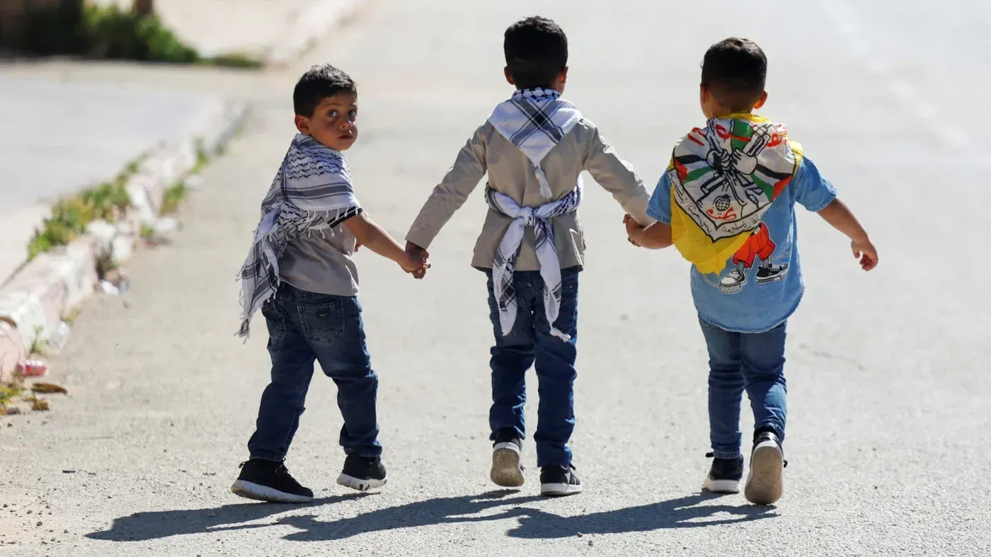 ثلاثة أطفال فلسطينيين يمسكون بأيدي بعضهم، يرتدون شالات تحمل رموزًا وطنية، يسيرون في شارع ضيق في ذكرى النكبة، معبرين عن الهوية الفلسطينية.