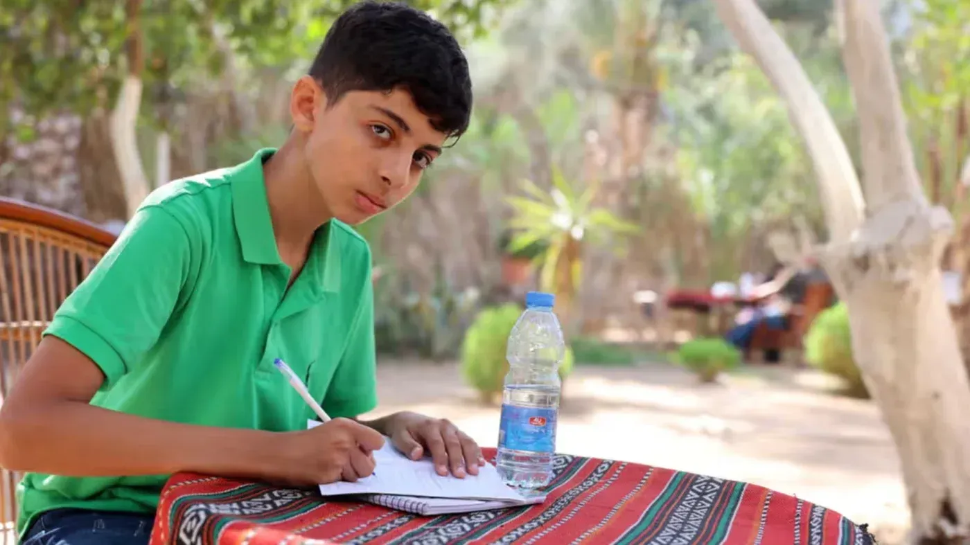 صبي يبلغ من العمر 13 عامًا يجلس على طاولة في حديقة، يكتب في دفتر ملاحظات، مع زجاجة ماء بجانبه.