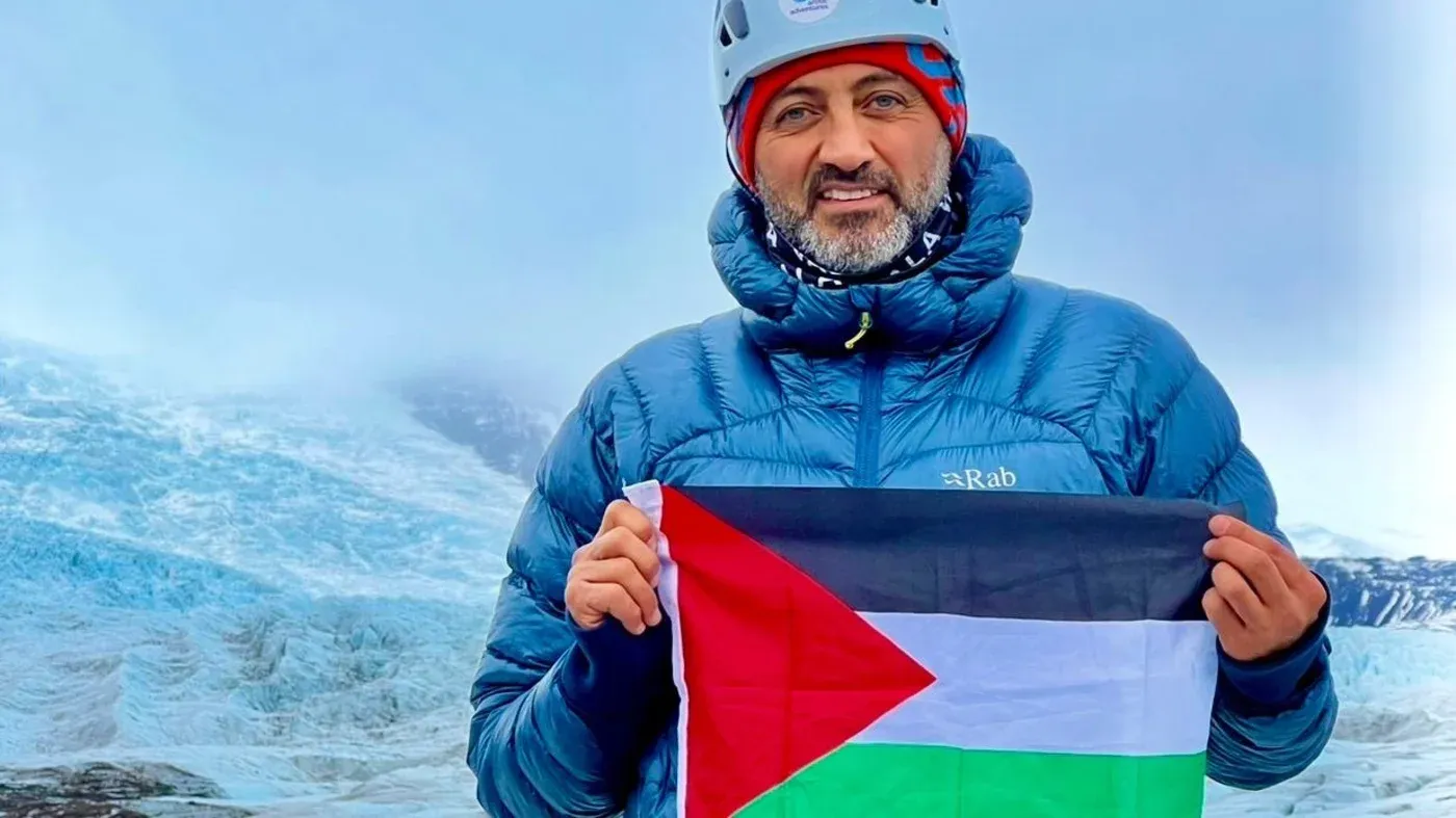 مصطفى سلامة يحمل علم فلسطين على جبل إيفرست، مرتديًا ملابس تسلق، مع خلفية جليدية، معبرًا عن أمله في دعم الأطفال في غزة.
