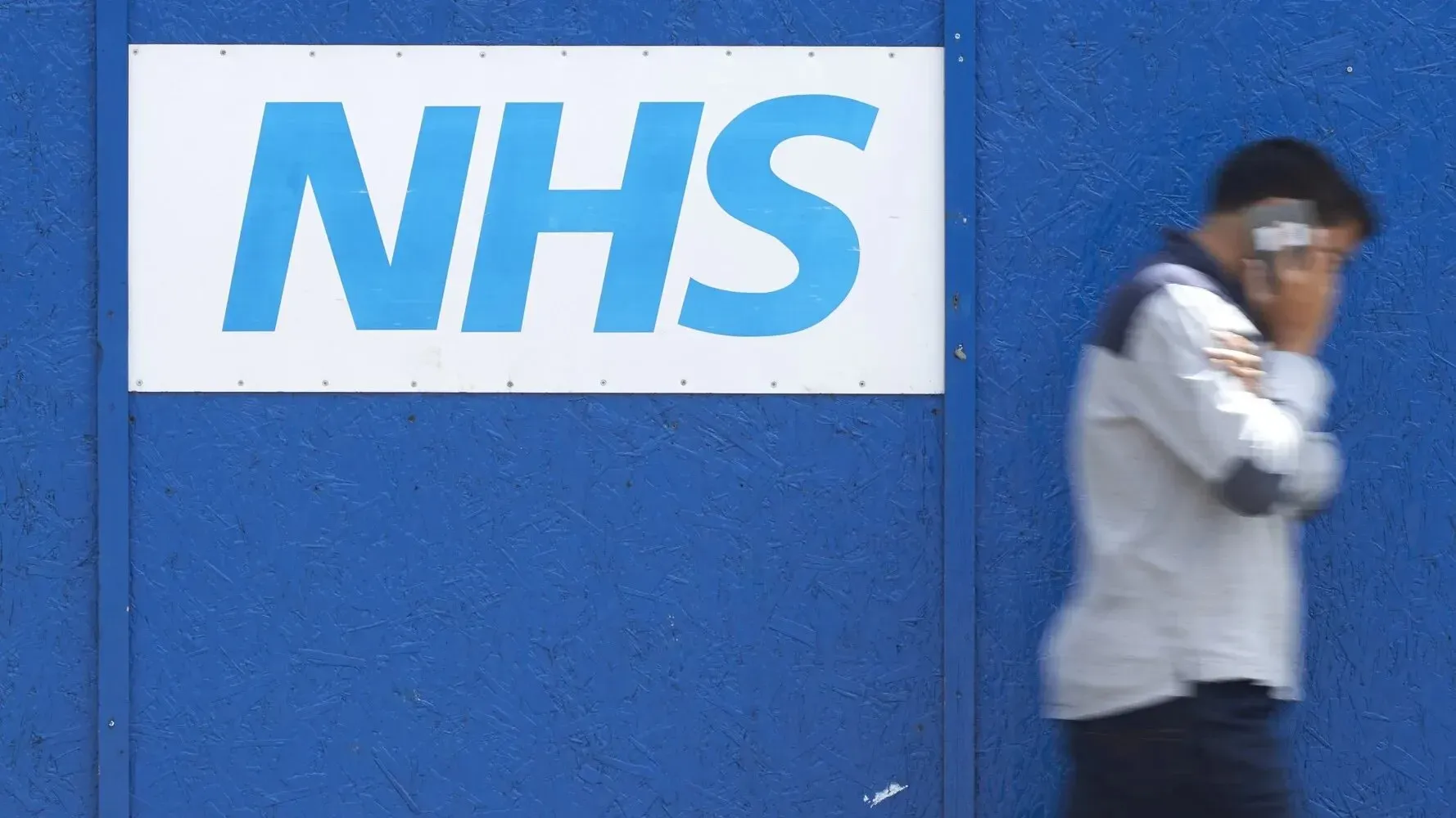 شخص يتحدث عبر الهاتف بجانب لافتة كبيرة تحمل شعار هيئة الخدمات الصحية الوطنية (NHS) على خلفية زرقاء.