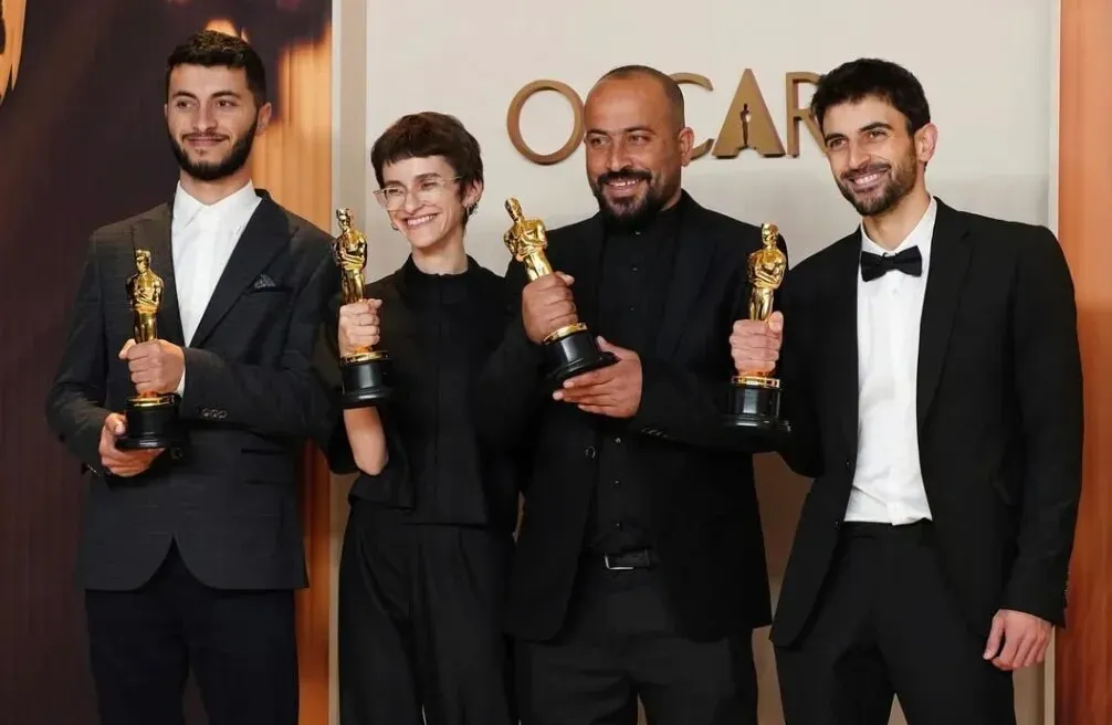 مخرج فلسطيني يحمل أوسكار، يقف مع زملائه في حفل توزيع الجوائز، مع تعبيرات فخرية، وسط خلفية احتفالية.