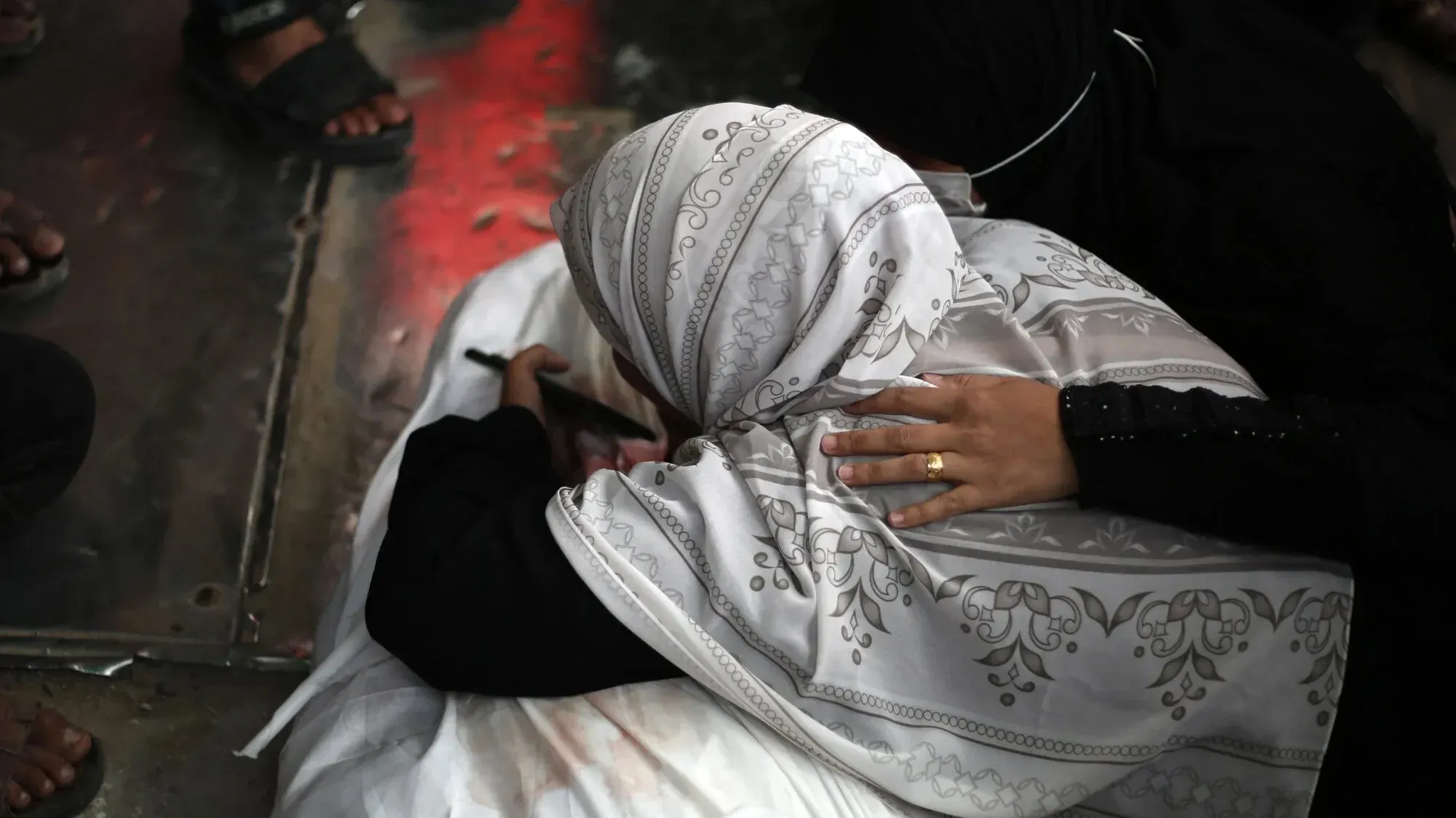امرأة ترتدي حجابًا أبيض، تجلس بجانب جثمان، تعبر عن الحزن بينما تضع يدها على الجثة، في مشهد يعكس الألم والمعاناة في غزة.