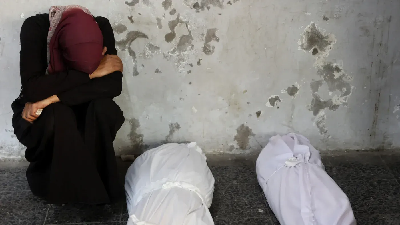 امرأة فلسطينية تجلس بجوار جثتين مغطاتين بالأقمشة البيضاء، تعبر عن الحزن والألم في خلفية مدمرة، تعكس معاناة المدنيين في غزة.