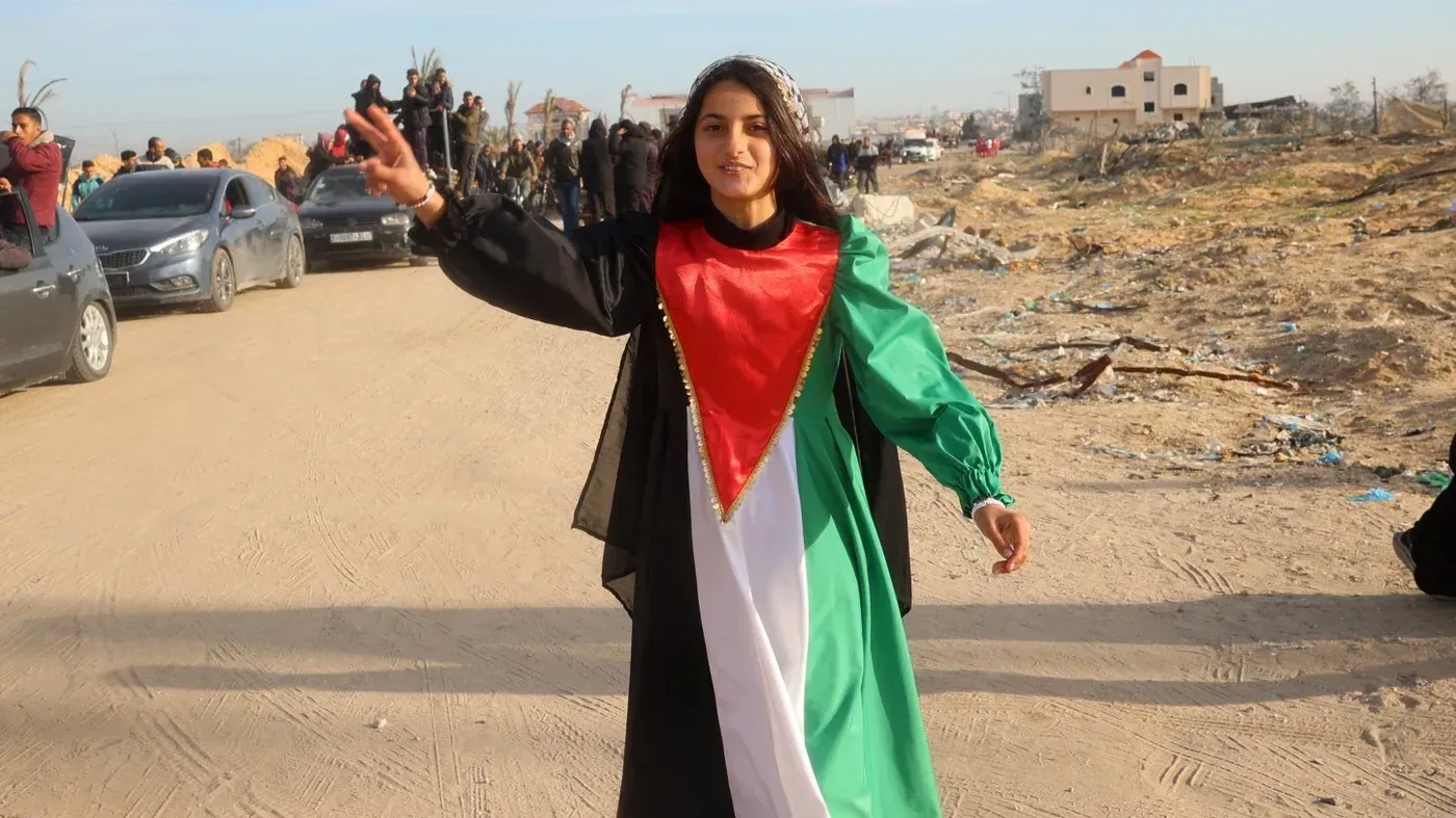 امرأة فلسطينية ترتدي زيًا تقليديًا يحمل ألوان العلم الفلسطيني، تتوسط مجموعة من السيارات في منطقة مدمرة، تعبر عن الأمل والمقاومة.