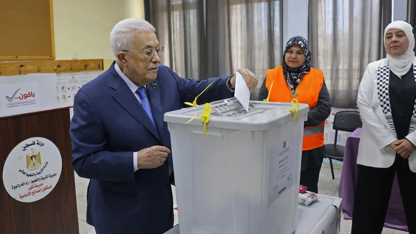 رجل مسن يُدلي بصوته في صندوق الاقتراع خلال الانتخابات البلدية الفلسطينية، بينما تراقبه امرأتان ترتديان زيًا رسميًا.