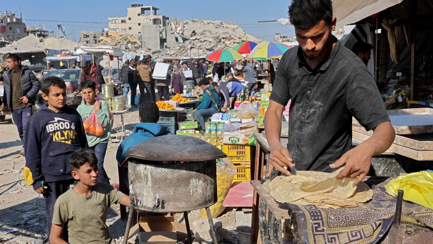 شخص يعمل على إعداد الخبز في سوق مزدحم في غزة، حيث تظهر خلفه أنقاض المباني المدمرة وأشخاص يتسوقون في ظروف صعبة.