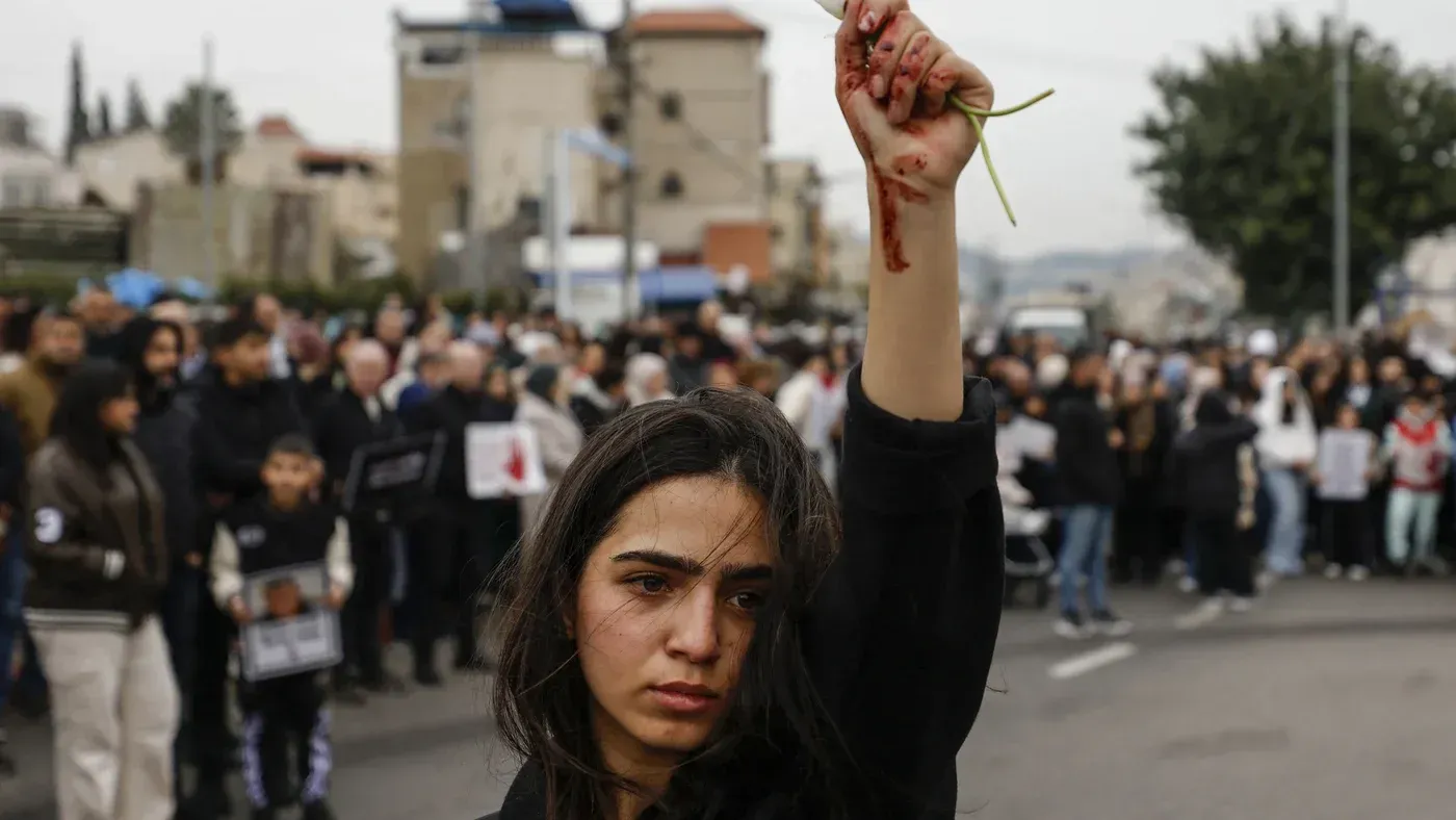 امرأة فلسطينية ترفع يدها عالياً وهي تحمل غصنًا أخضر، بينما تظهر على وجهها علامات الدم، وسط حشد من المتظاهرين في الخلفية.