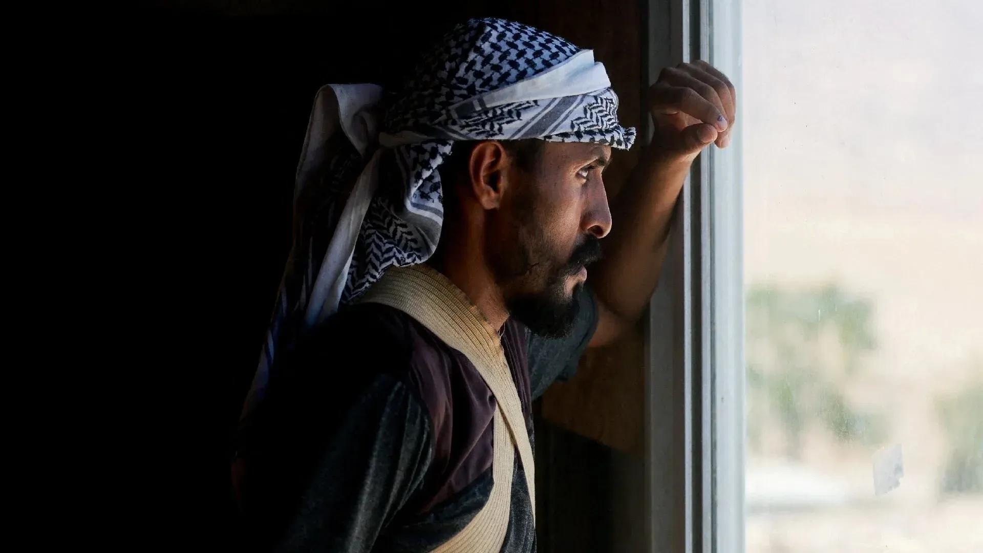 رجل فلسطيني يرتدي الكوفية، يراقب من نافذة منزله في قرية بدوية بالضفة الغربية، معبرًا عن القلق بسبب تصاعد العنف من المستوطنين.