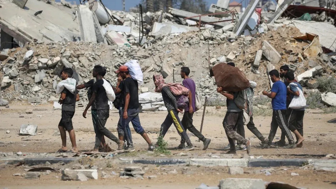 مجموعة من الفلسطينيين يسيرون وسط أنقاض المباني المدمرة في غزة، يحملون حقائب وأمتعة، في سياق أزمة إنسانية متزايدة.