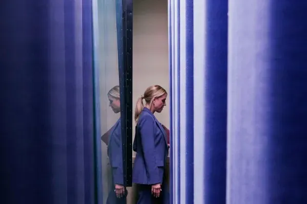 امرأة ترتدي بدلة زرقاء تقف بين عمودين زرقاوين، تعكس صورتها في الزجاج، مما يرمز إلى التوتر في وزارة العدل خلال فترة بام بوندي.