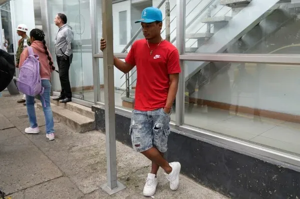 شاب يرتدي قميصًا أحمر وقبعة زرقاء، يستند إلى عمود في منطقة حضرية، بينما يقف آخرون في الخلفية، مما يعكس الحياة اليومية في كولومبيا.