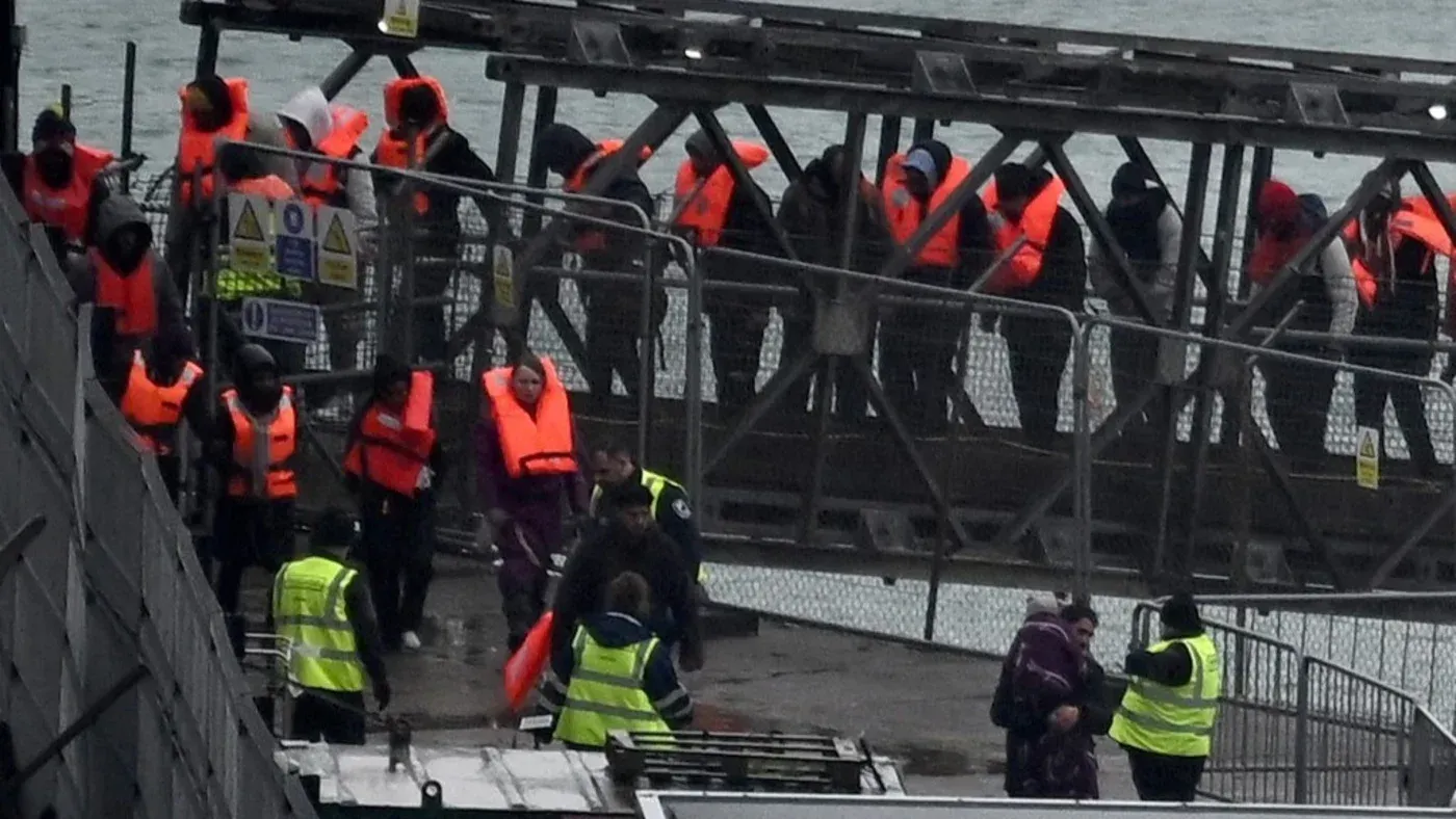 طالبي اللجوء يرتدون سترات نجاة برتقالية أثناء وصولهم إلى المملكة المتحدة عبر قوارب صغيرة، وسط إجراءات أمنية مشددة.