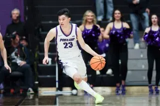 لاعب كرة سلة من فريق بورتلاند يحمل الكرة أثناء المباراة، مع وجود مشجعين في الخلفية يشجعون الفريق.