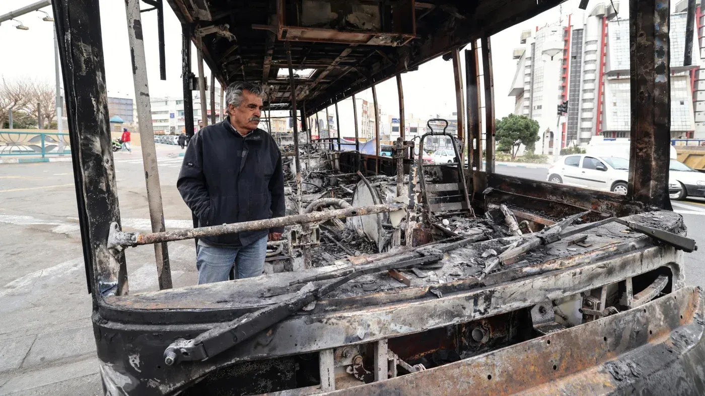 رجل يقف بجانب حافلة محترقة في إيران، تعكس آثار الاضطرابات والاحتجاجات المستمرة، وسط تزايد التوترات السياسية.