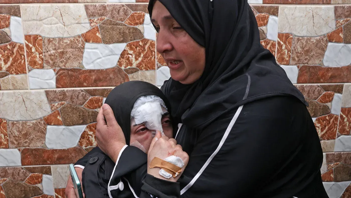امرأة ترتدي الحجاب تحتضن طفلة مصابة، بينما تظهر علامات الحزن على وجههما، في خلفية جدارية ملونة، بعد هجوم صاروخي في الضفة الغربية.