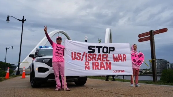 ناشطون يرتدون ملابس وردية يحملون لافتة تطالب بوقف الحرب الأمريكية الإسرائيلية على إيران، بينما تقف سيارة شرطة في الخلفية.