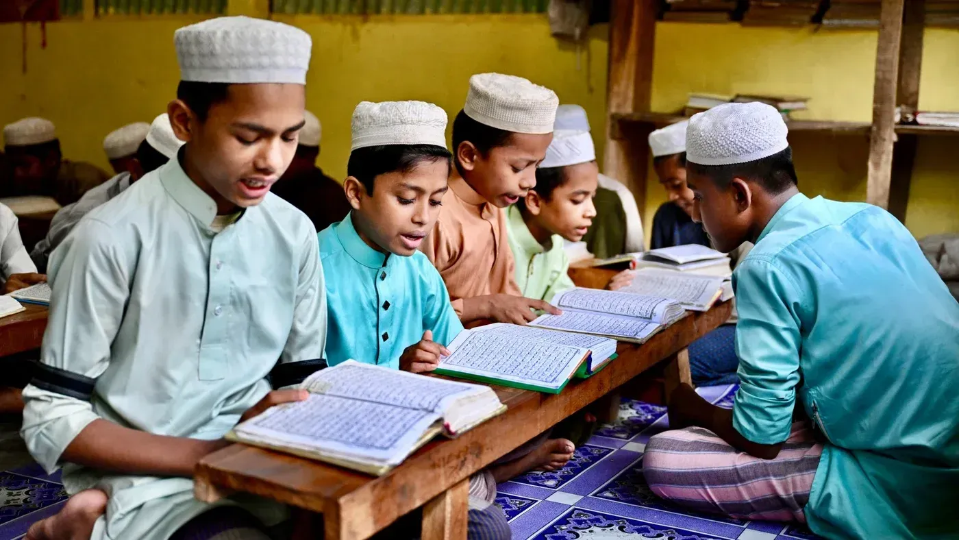 مجموعة من الأطفال يرتدون قمصانًا تقليدية وقبعات، يقرأون القرآن في فصل دراسي، مما يعكس أهمية التعليم الديني خلال شهر رمضان.