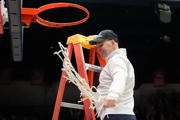 مدرب كرة السلة راندي بينيت يقف على سلم ويقطع شبكة السلة احتفالًا بفوز فريقه، مما يعكس نجاحه في مسيرته التدريبية.