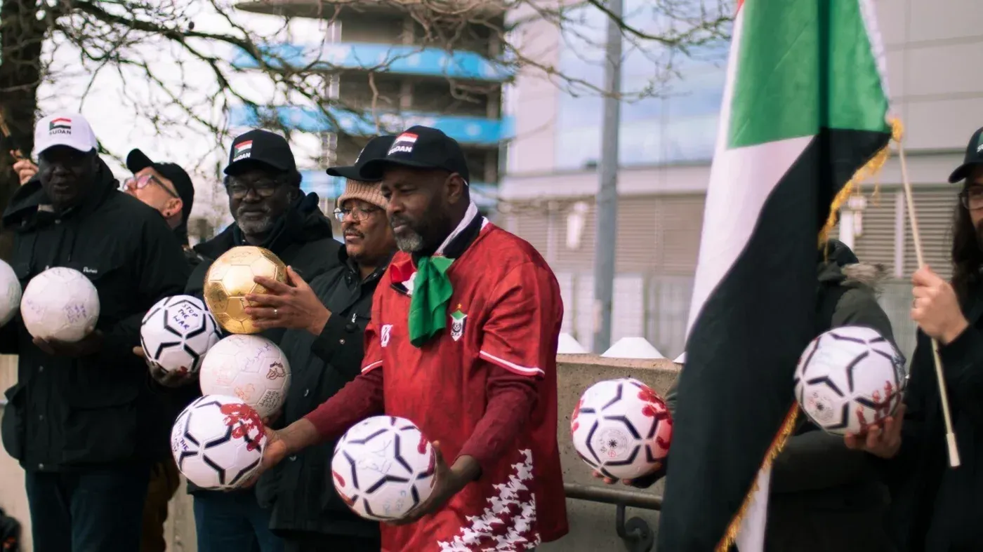 محتجون سودانيون أمام ملعب مانشستر سيتي يحملون كرات قدم وأعلام، يطالبون بوقف دعم الشيخ منصور لقوات الدعم السريع في السودان.