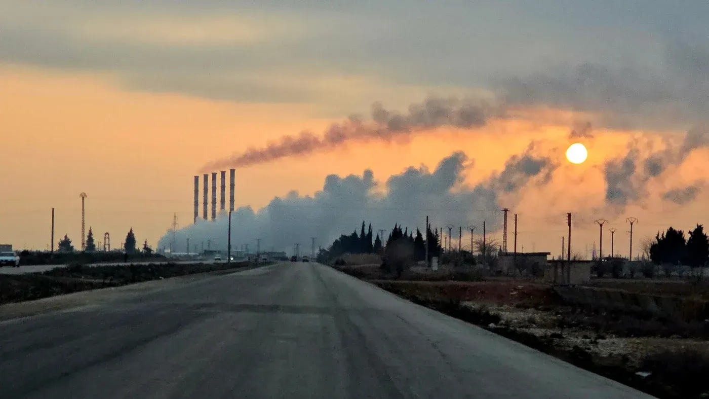 غروب الشمس خلف مصانع في شمال شرق سوريا، مع تصاعد الدخان في السماء، يعكس التحديات البيئية والاقتصادية في المنطقة.