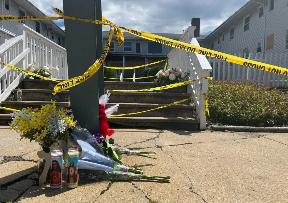 موقع حادث حريق منزل غابرييل هاوس في فال ريفر، مع زهور وشموع تذكارية، وشريط تحذيري يحيط بالمكان.
