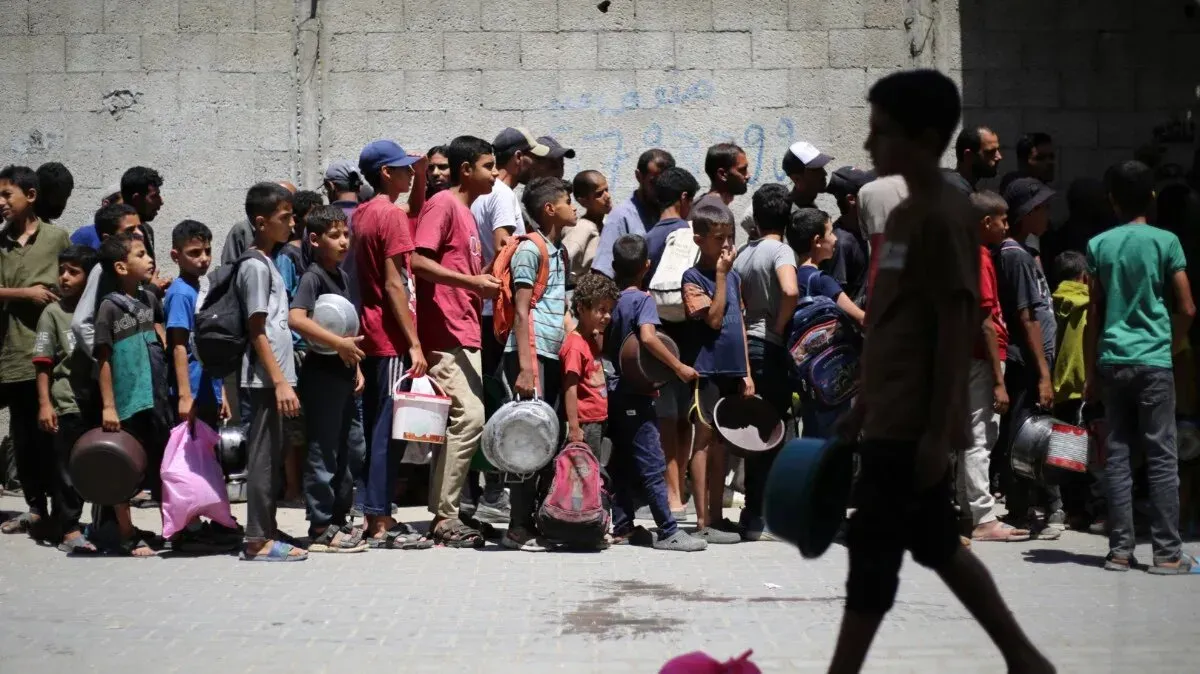 طابور طويل من الأطفال والبالغين في غزة يحملون أواني وأكياس، ينتظرون للحصول على المساعدات الإنسانية وسط الدمار.