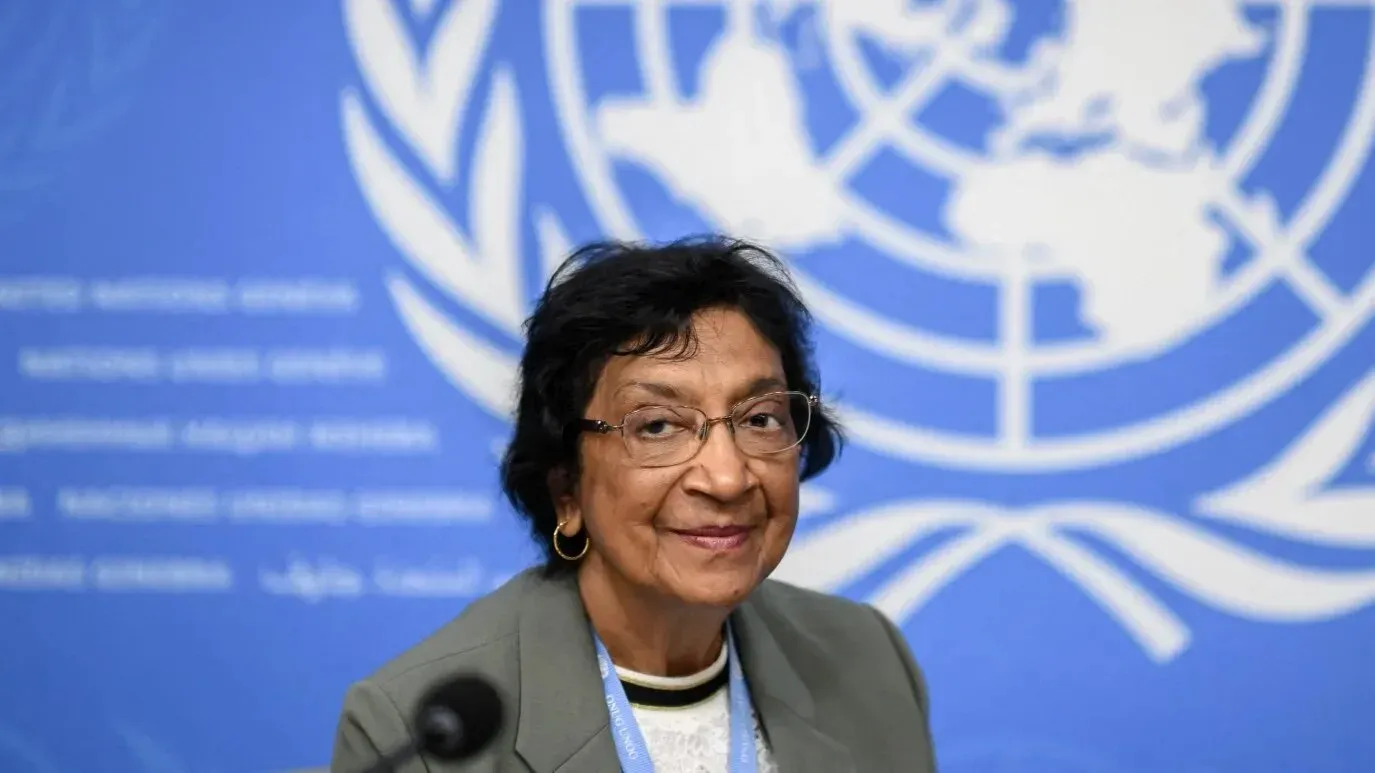 صورة لنافي بيلاي، رئيسة لجنة التحقيق التابعة للأمم المتحدة بشأن فلسطين، مبتسمة أمام خلفية شعار الأمم المتحدة.