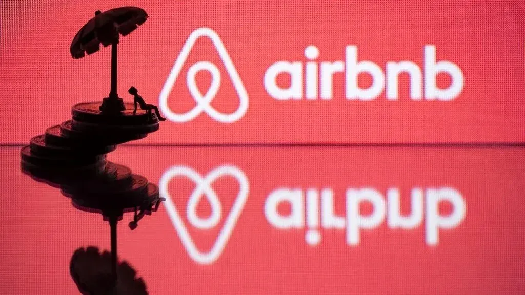 شعار شركة Airbnb مع مجسم لشخص يجلس تحت مظلة، يعكس القضايا القانونية المتعلقة بممارسات الشركة في المستوطنات الإسرائيلية.