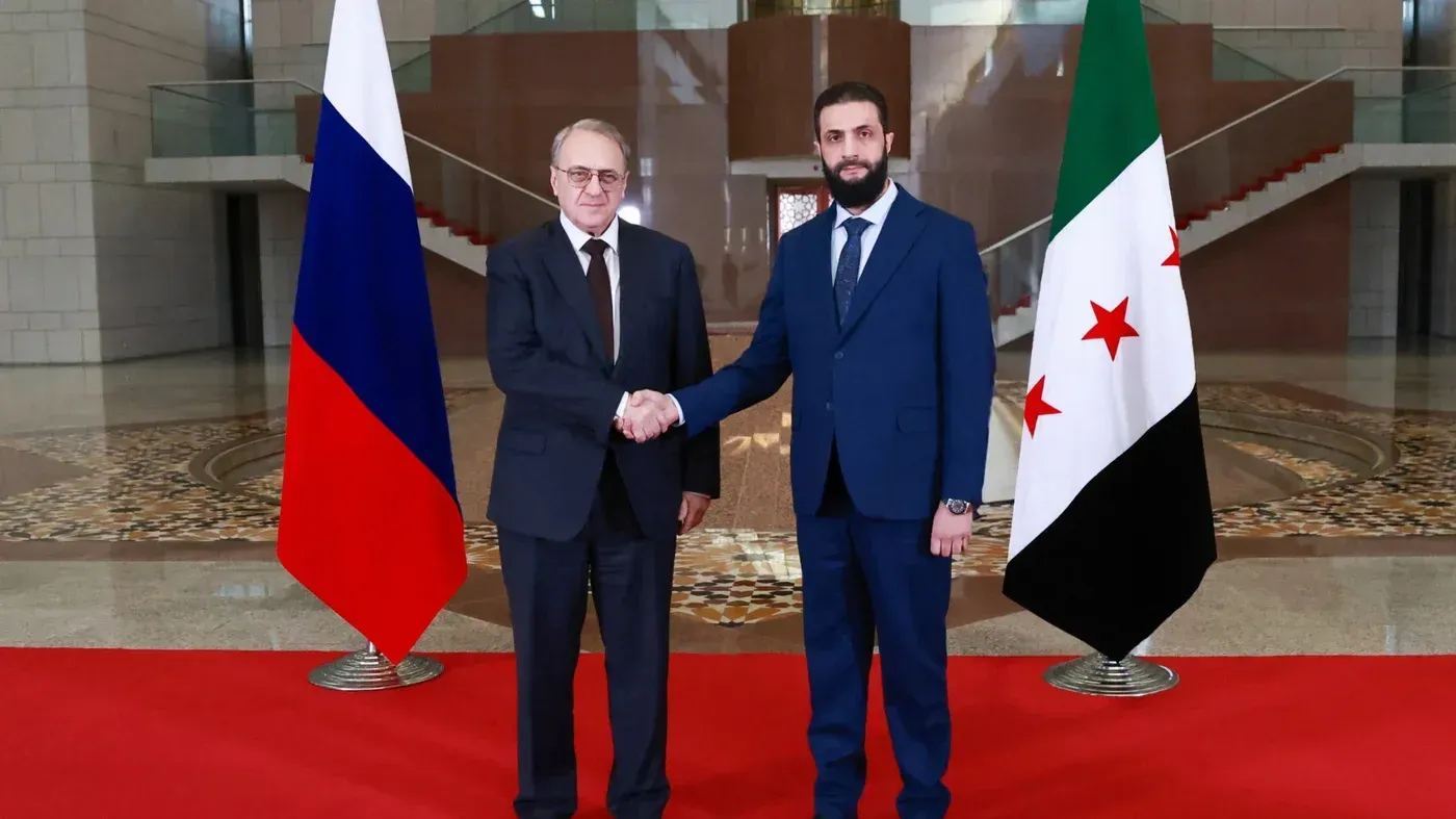 اجتماع بين مسؤول روسي وقيادي سوري، مع العلم الوطني الروسي والسوري خلفهما، يعكس العلاقات المتنامية بين البلدين.