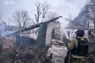 رجل إطفاء يرتدي سترة واقية يرش المياه على حطام مبنى مدمر، مع دخان يتصاعد من الأنقاض وأشجار عارية في الخلفية، مما يعكس آثار القصف في أوكرانيا.