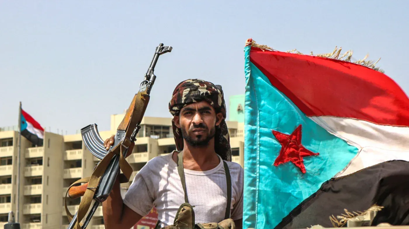 مقاتل من المجلس الانتقالي الجنوبي يحمل سلاحه أمام علم اليمن الجنوبي، في سياق تصاعد التوترات بين السعودية والإمارات.