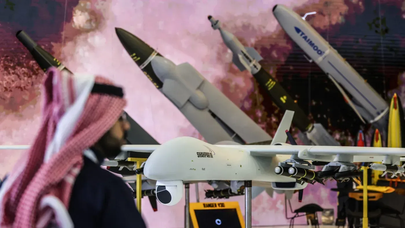 رجل يرتدي الزي السعودي يقف أمام طائرات مسيرة وصواريخ في معرض عسكري، مما يعكس التوترات الإقليمية والجهود الدفاعية في الخليج.