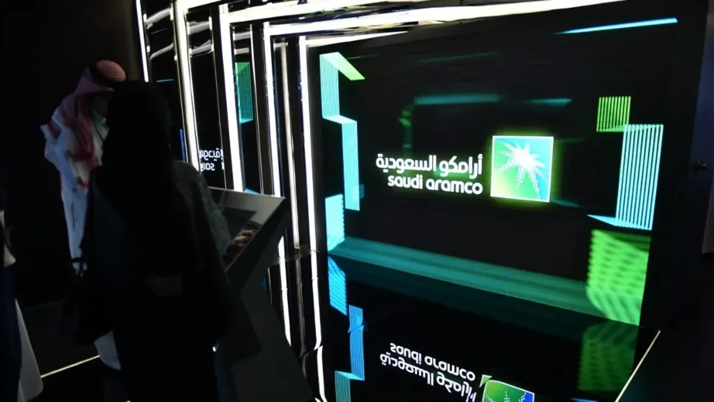 شخصية تتفاعل مع عرض رقمي لشعار أرامكو السعودية في بيئة حديثة، تعكس التحديات الاقتصادية الحالية للشركة.