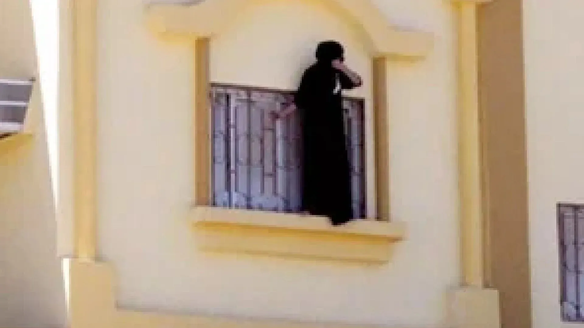امرأة ترتدي العباءة وتقف على حافة نافذة في دار رعاية، تعبر عن شعورها بالاحتجاز والخوف من الظروف القاسية التي تواجهها.